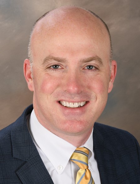 A person with a shaved head wearing a white shirt and a blue striped blazer against a brown background.