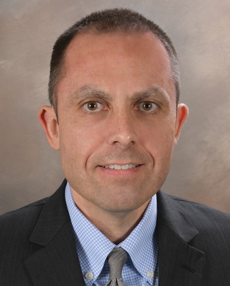 A person with short dark brown hair wearing a blue shirt with a dark grey blazer against a brown background.