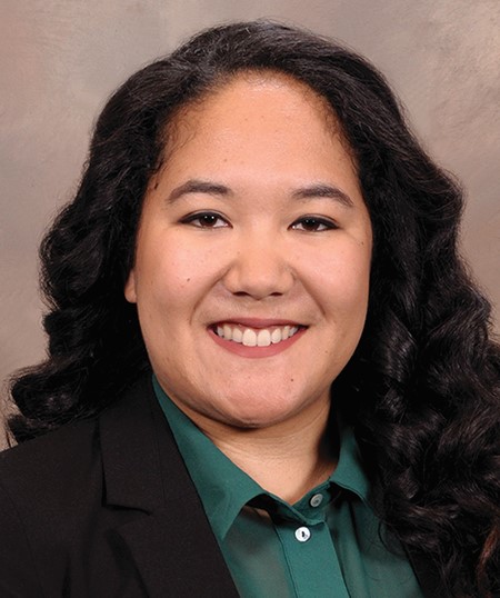 A person with long black hair wearing a green shirt with a black blazer against a brown background.