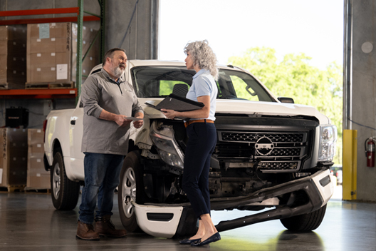 Two individuals conversing in front of a damaged white truck.