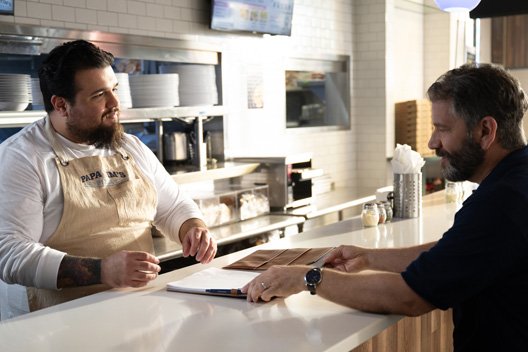 An individual behind a counter wearing an apron talking to another individual holding a notebook.